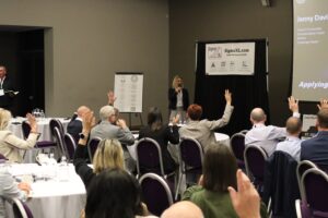 Jenny Davis Lean Six Sigma ILSSI Conference 2025 Jersey with audience hands up using Lean Six Sigma for coping with and fighting and winning against cancer